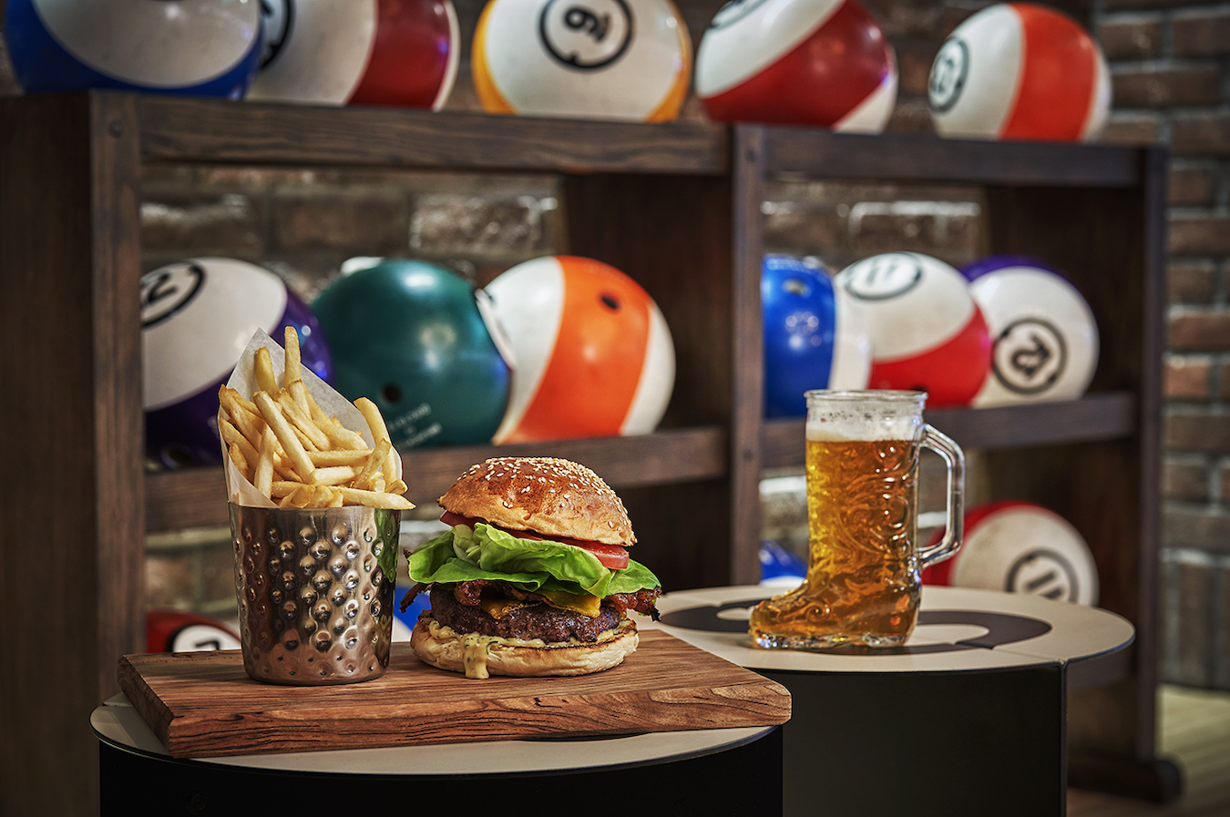 A burger with lettuce, tomato, and bacon on a wooden board sits next to a metal container of fries. A glass boot mug filled with beer is on the right. The background features shelves with colorful billiard balls.