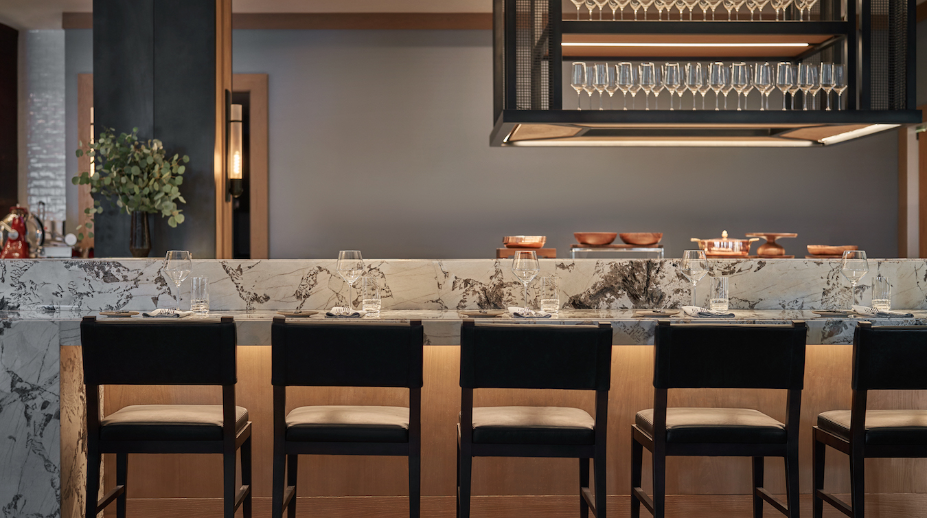 A modern restaurant bar with a marble countertop. Four black cushioned chairs are neatly arranged along the counter. Above, rows of upside-down wine glasses hang from a rack. The counter is set with napkins, glasses, and small plates, with soft, ambient lighting.