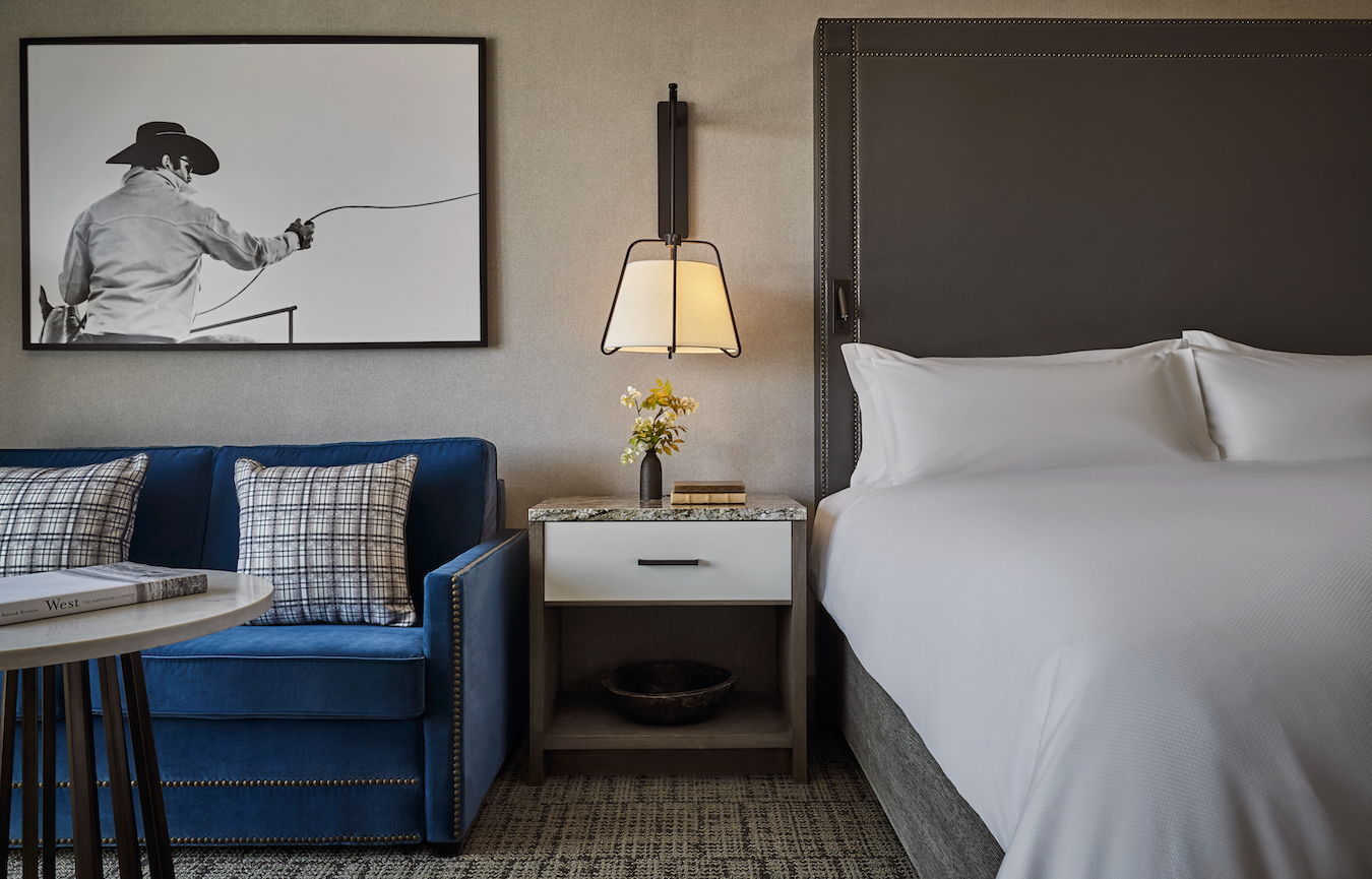 A modern hotel room featuring a plush, white bed, a nightstand with a lit lamp, and a vase of yellow flowers. To the left, there's a blue sofa with plaid pillows. Above the sofa hangs a black and white photo of a cowboy with a lasso. The decor is contemporary.