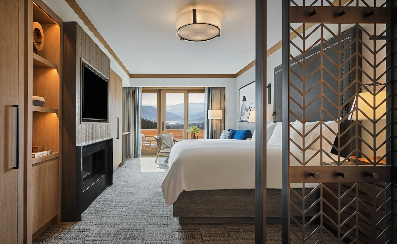 A modern hotel room features a queen-sized bed with white linens, a wood-paneled television, and open shelves on the left. A sliding partition partially reveals a cozy seating area by a large window offering a scenic mountain view. Neutral colors and sleek décor.