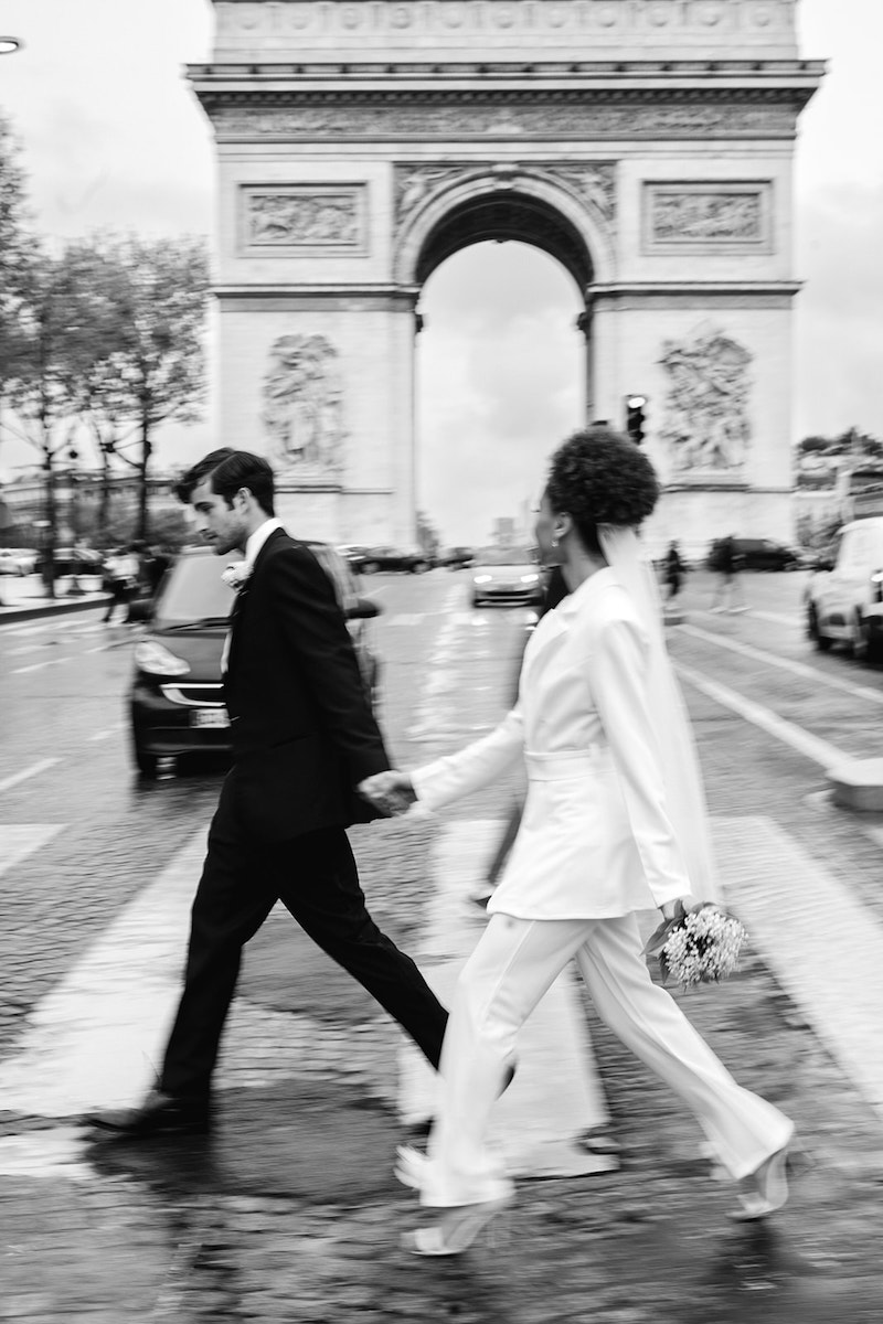 A couple dressed in a wedding suit and a bridal outfit cross a street in front of the Arc de Triomphe on a rainy day. The bride holds a small bouquet of flowers.