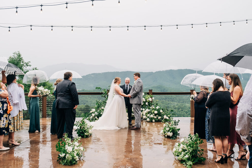 A bride and groom stand at an outdoor wedding ceremony under string lights, holding hands, while guests observe with umbrellas on a rainy day. The background shows a foggy mountain landscape.