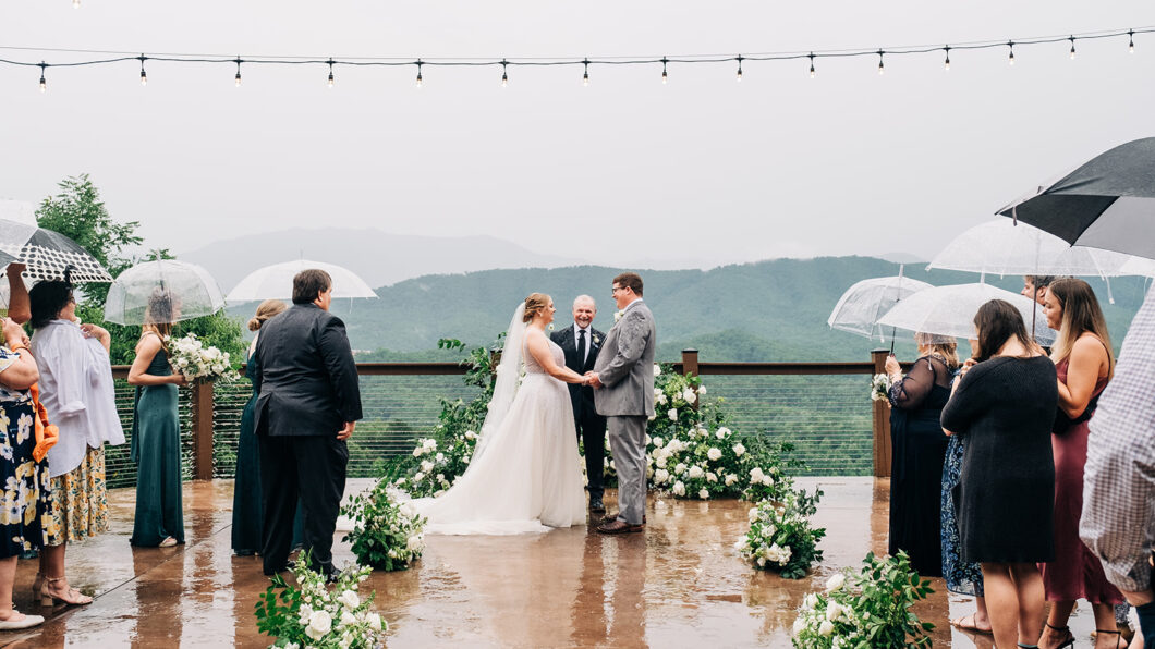 A bride and groom stand at an outdoor wedding ceremony under string lights, holding hands, while guests observe with umbrellas on a rainy day. The background shows a foggy mountain landscape.