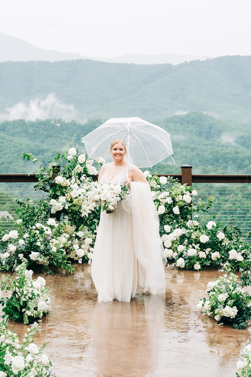 A bride in a white gown stands under a clear umbrella on a wet patio, surrounded by white flowers, with a mountain landscape in the background.