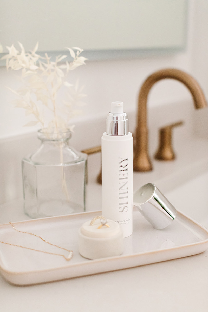 Bottle of Shinery jewelry cleaner on a bathroom counter with gold fixtures, beside a vase with dried plants and jewelry displayed on a tray.