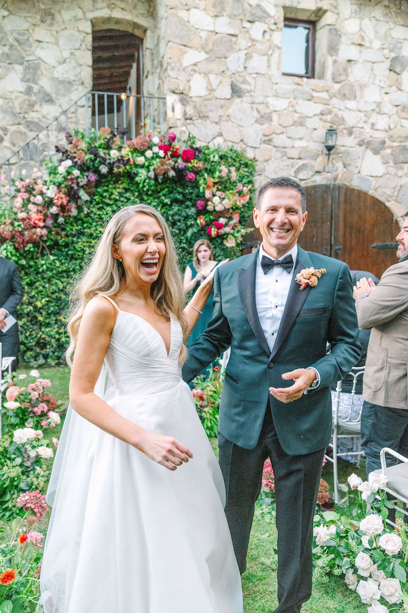 A bride in a white dress and a man in a tuxedo stand smiling in front of a stone building adorned with flowers.