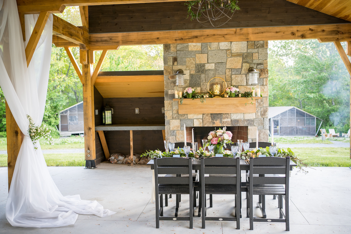 An outdoor dining area under a wooden pavilion, featuring a table set for six with floral centerpieces. A stone fireplace is in the background, and white drapes hang on the pavilion's side.