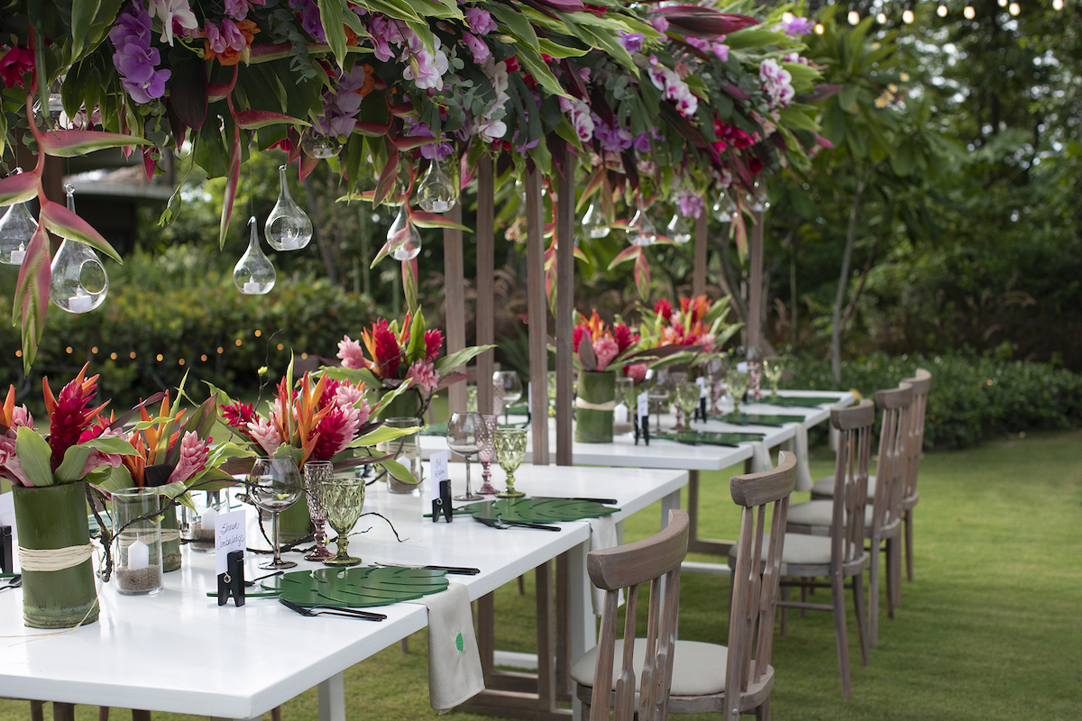 A long table set for an outdoor event with vibrant floral arrangements, hanging glass decorations, and wooden chairs, situated in a lush garden setting.