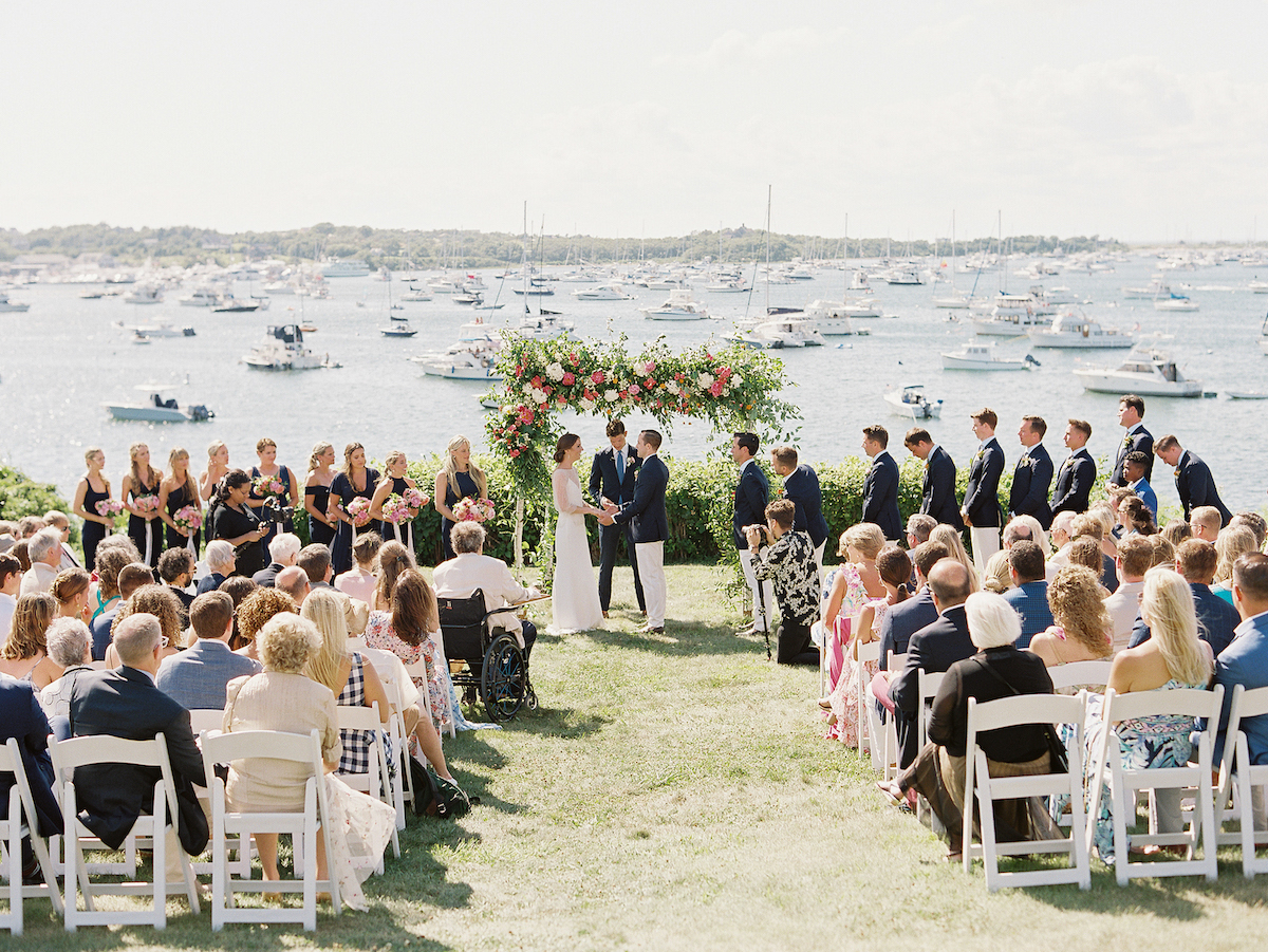 A couple stands under a floral arch during an outdoor wedding ceremony by the water, with bridesmaids and groomsmen on either side and guests seated in rows facing the couple.