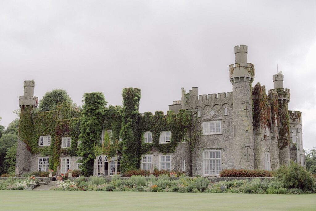 A large stone castle covered in ivy with multiple towers and windows, surrounded by a neatly maintained garden and expansive lawn. The sky above is overcast.