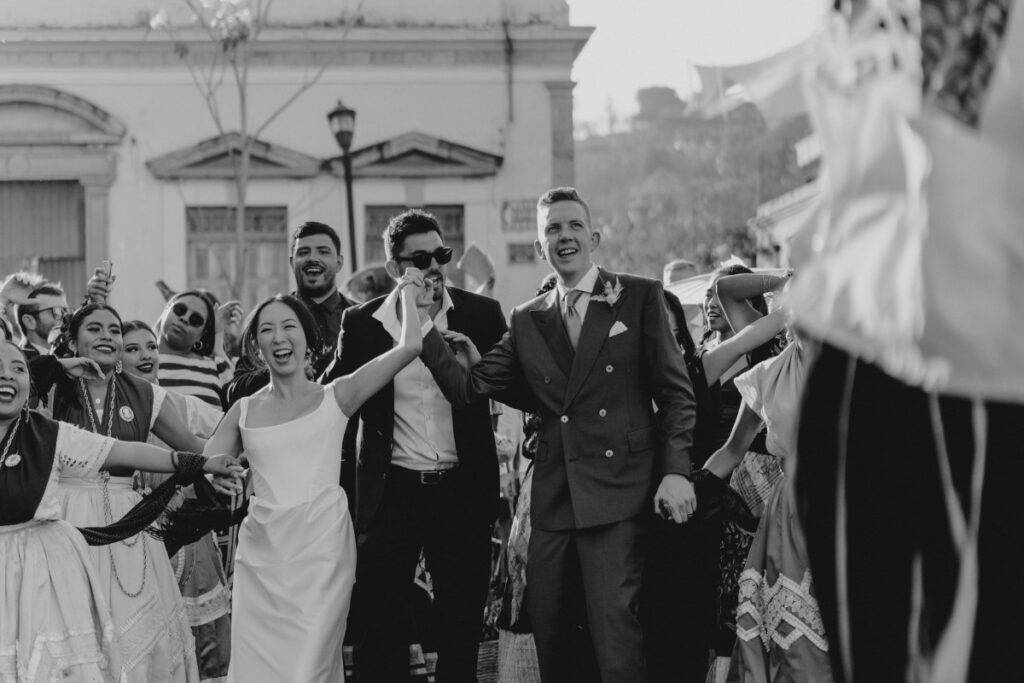 A black and white image of a wedding procession with a joyful bride and groom holding hands, surrounded by smiling, formally dressed guests and performers in traditional attire.