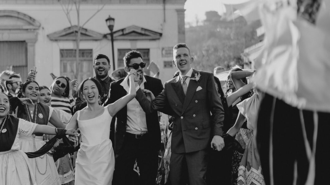 A black and white image of a wedding procession with a joyful bride and groom holding hands, surrounded by smiling, formally dressed guests and performers in traditional attire.