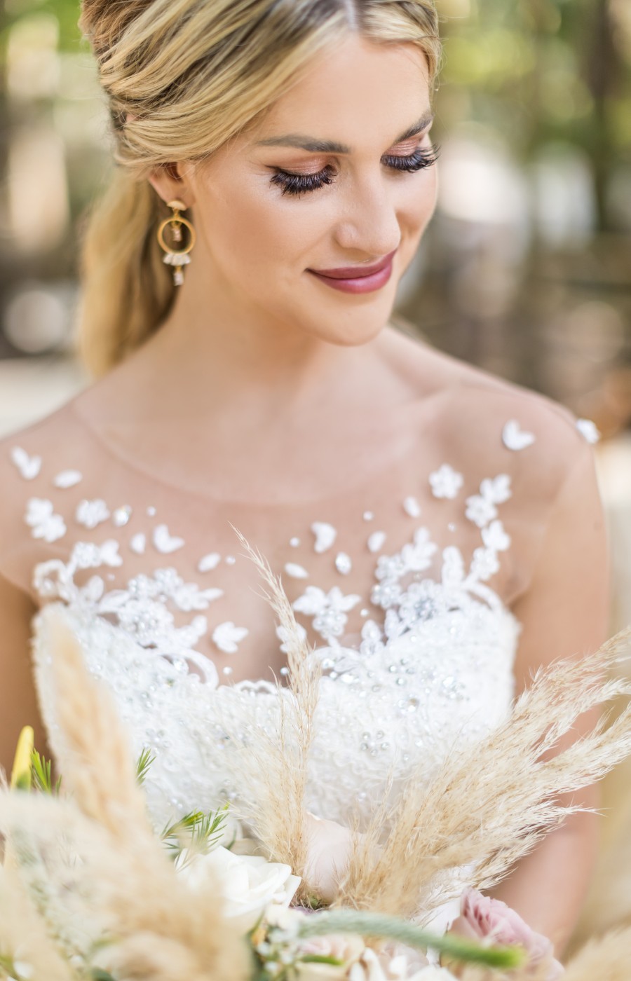 A bride in a white, floral-patterned wedding dress holds a bouquet and smiles softly, looking down. She has blond hair styled back and wears drop earrings. Trees are visible in the blurred background.