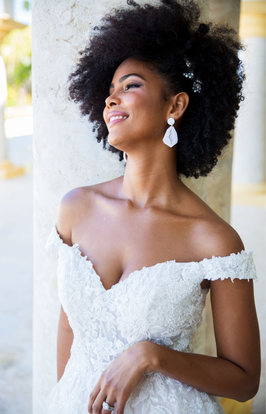 Bride in an off-shoulder lace wedding dress, smiling with a relaxed pose, leaning against a column. She has an afro hairstyle adorned with small flowers and wears white earrings.