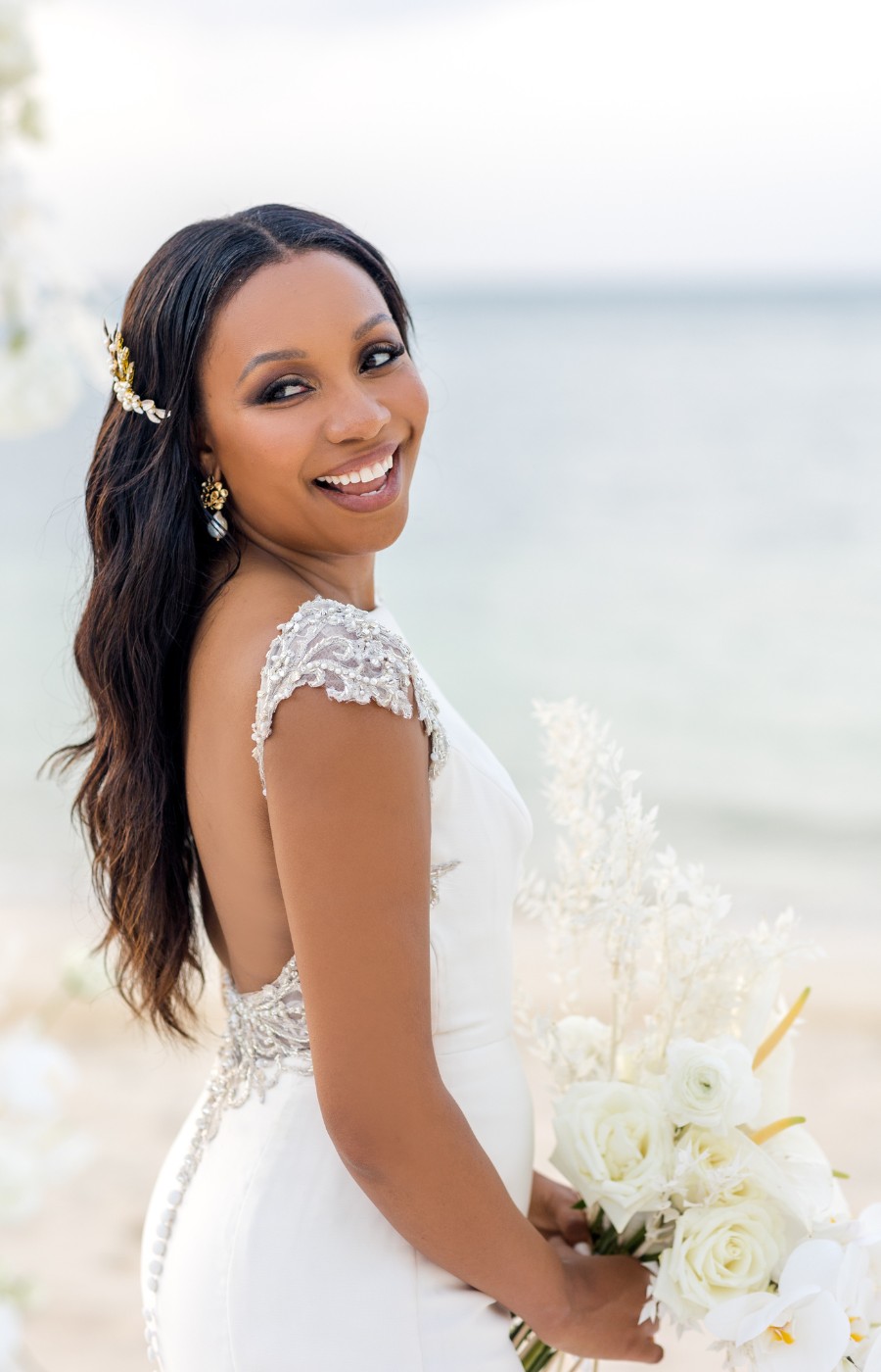 A bride in a white dress with lace details smiles while holding a bouquet of white flowers in front of a beach backdrop.