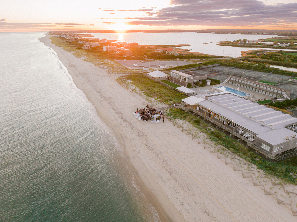 Aerial view of a wedding ceremony taking place on a sandy beach near a coastal resort at sunset. The setup includes chairs arranged in front of a small gazebo. The ocean is calm with gentle waves.