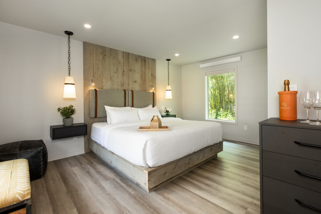 A modern bedroom with a large bed, wooden headboard, two hanging lamps, a potted plant, a black dresser with an orange bucket, and a window showing green foliage outside.