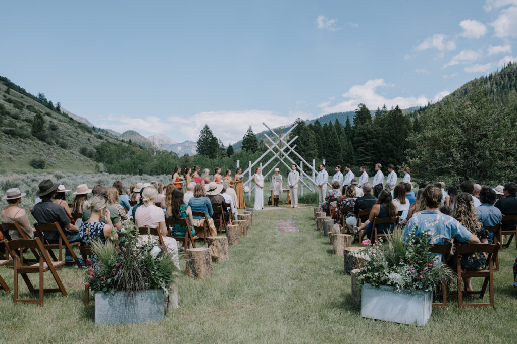 Outdoor wedding ceremony with guests seated, a wedding altar made of wooden sticks, and mountains in the background. The wedding party stands on either side of the couple in the center.