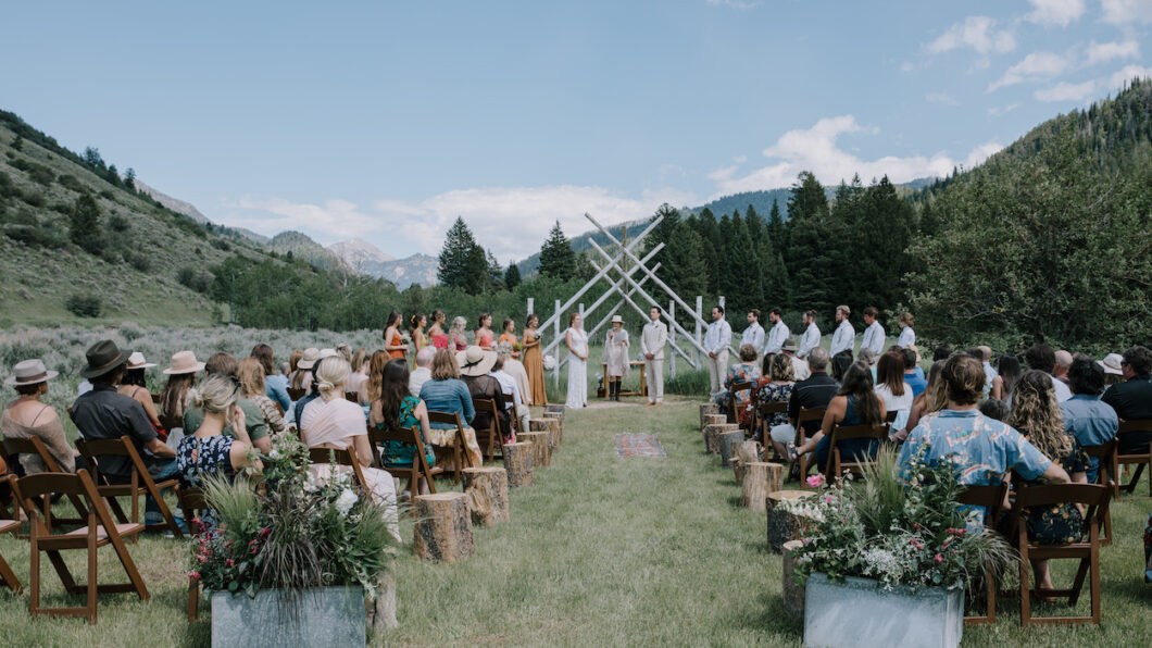 Outdoor wedding ceremony with guests seated, a wedding altar made of wooden sticks, and mountains in the background. The wedding party stands on either side of the couple in the center.