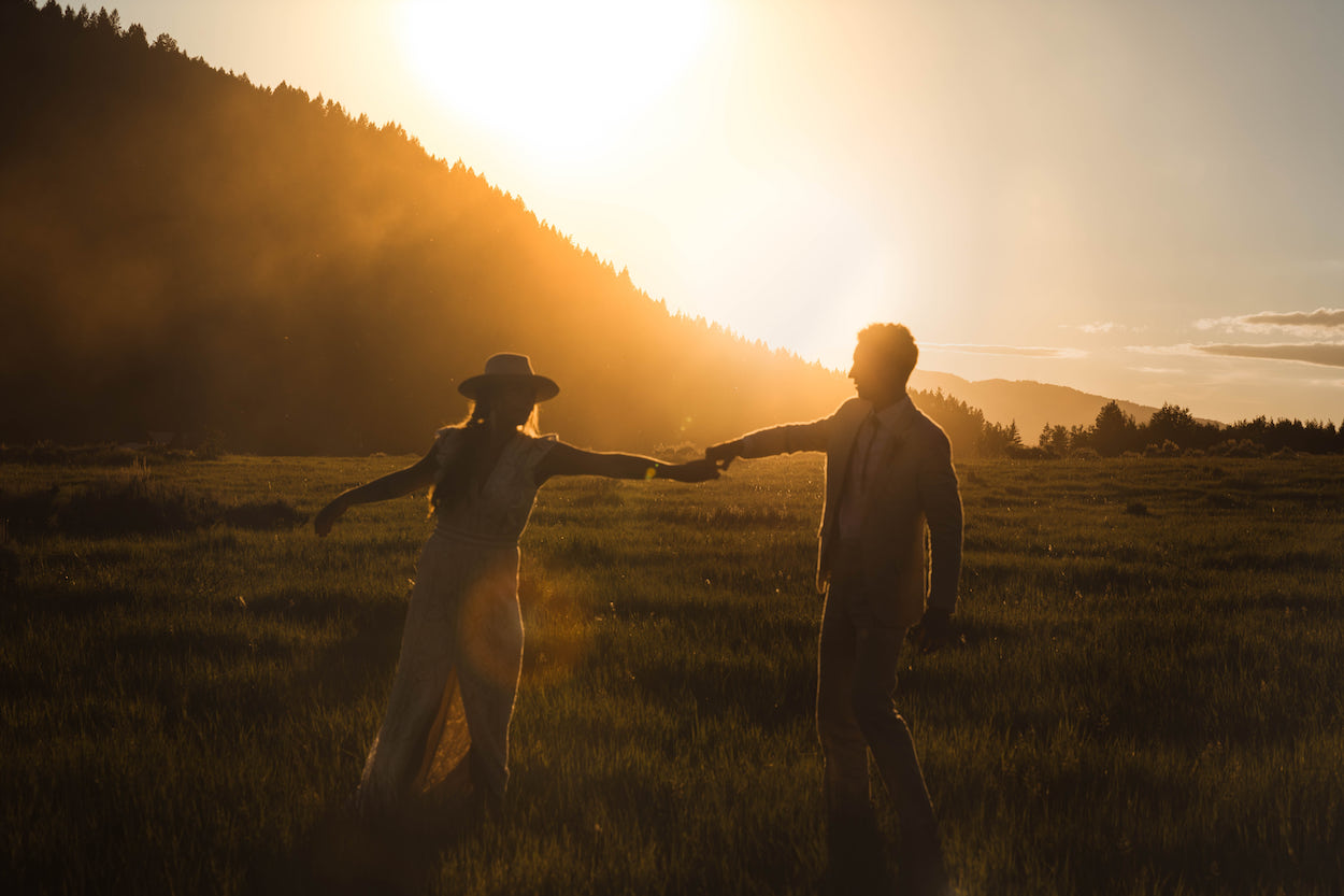 A couple holds hands and dances in a grassy field at sunset, with a mountainous landscape in the background.