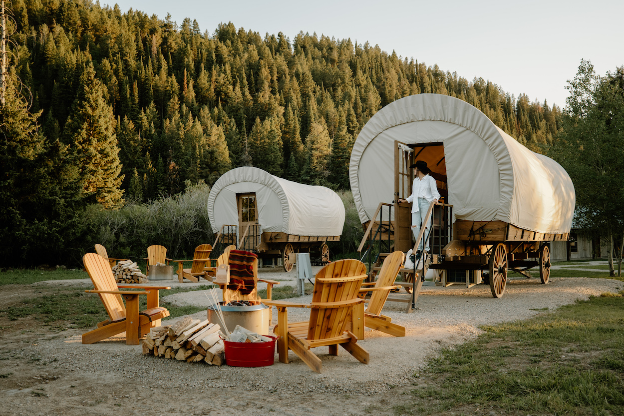 Two covered wagons used as cabins near a forest, with a person exiting one. Four wooden chairs surround a fire pit with a stack of firewood beside it.