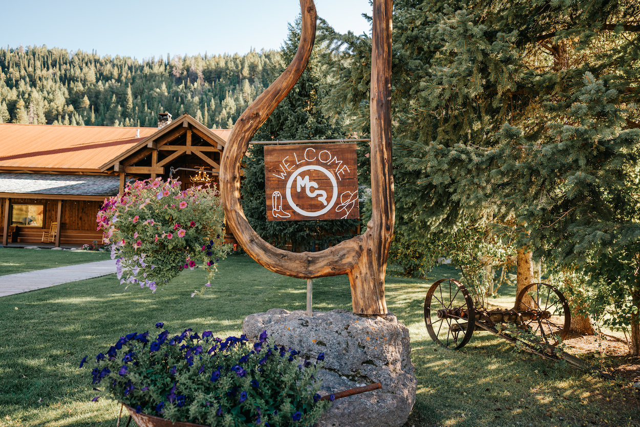 Wooden sign with "Welcome" and "MC" logo situated in a rustic outdoor setting with a wheelbarrow of flowers, an old wagon wheel, and a wooden building in the background.