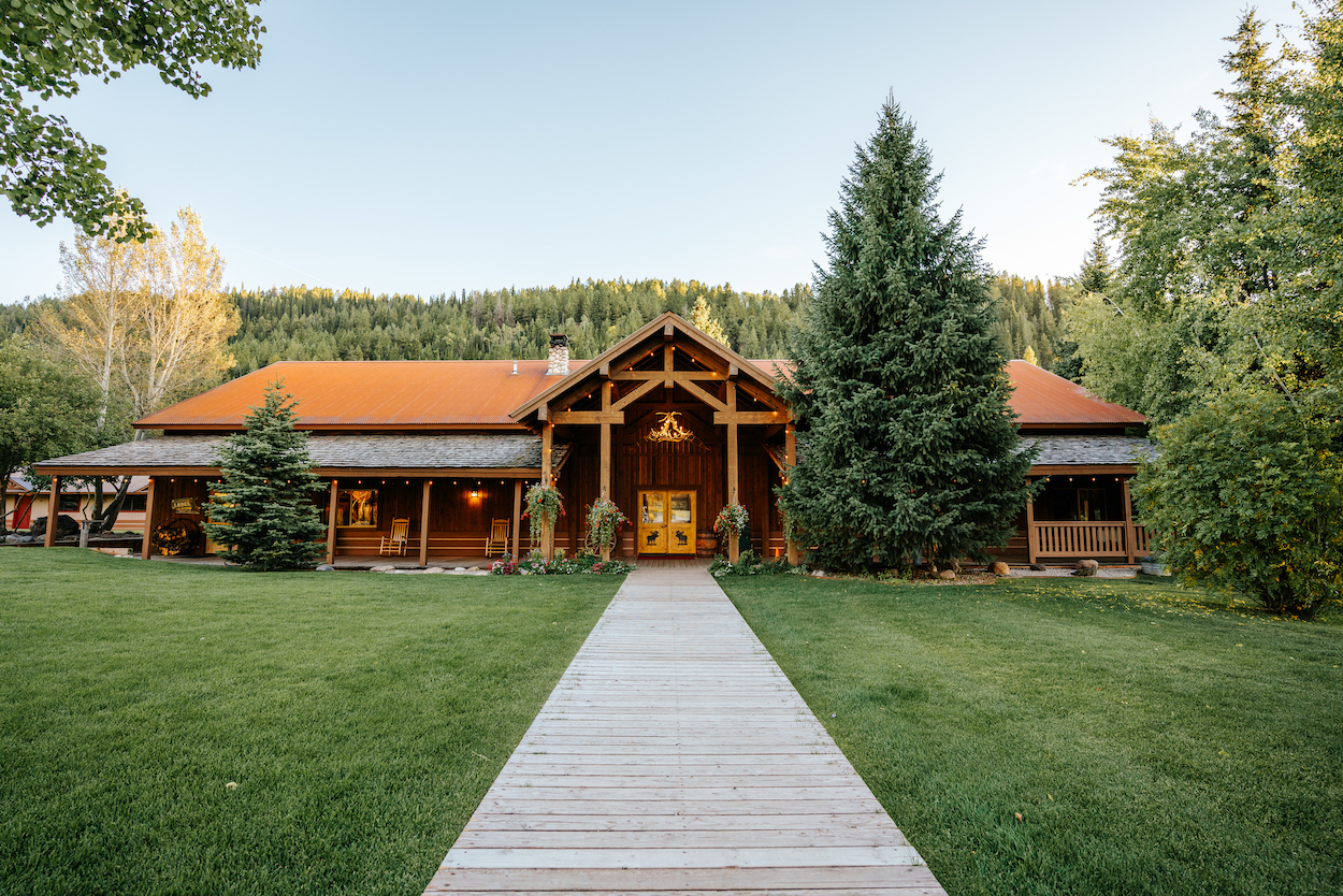 A large wooden lodge with a red roof sits amid green lawns and trees, with a pathway leading to the entrance.