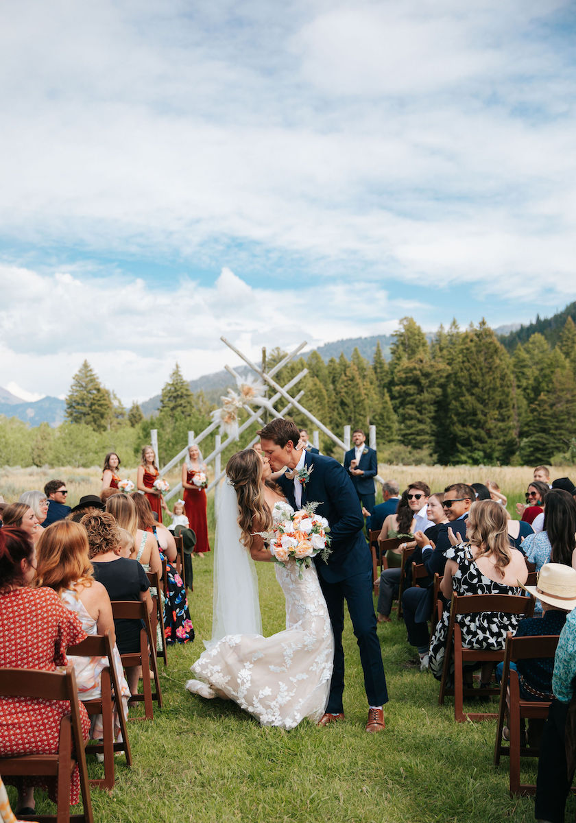 A bride and groom share a kiss at an outdoor wedding ceremony. Guests seated on either side in a grassy setting with trees and mountains in the background.