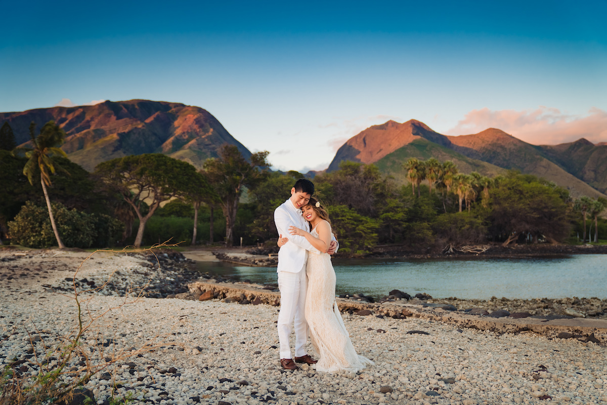 A couple in wedding attire embraces on a rocky beach with lush greenery and mountains in the background under a clear blue sky.