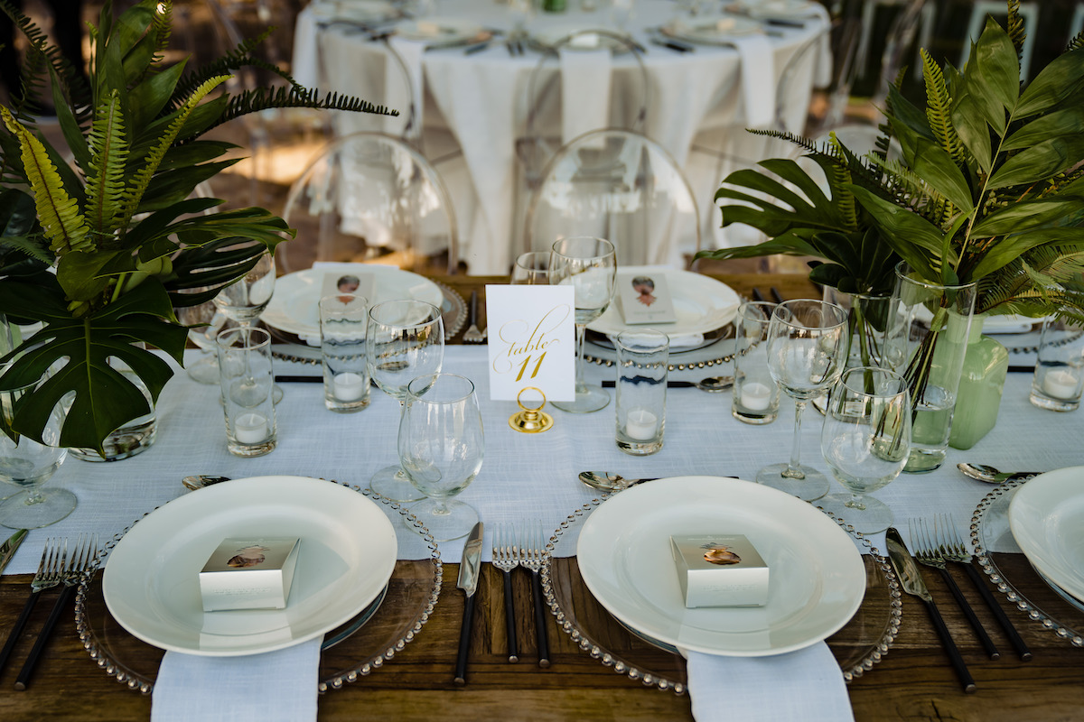 A dining table set for an event, featuring white plates, glassware, and green leafy centerpieces. A table number card showing "11" is placed in the center.