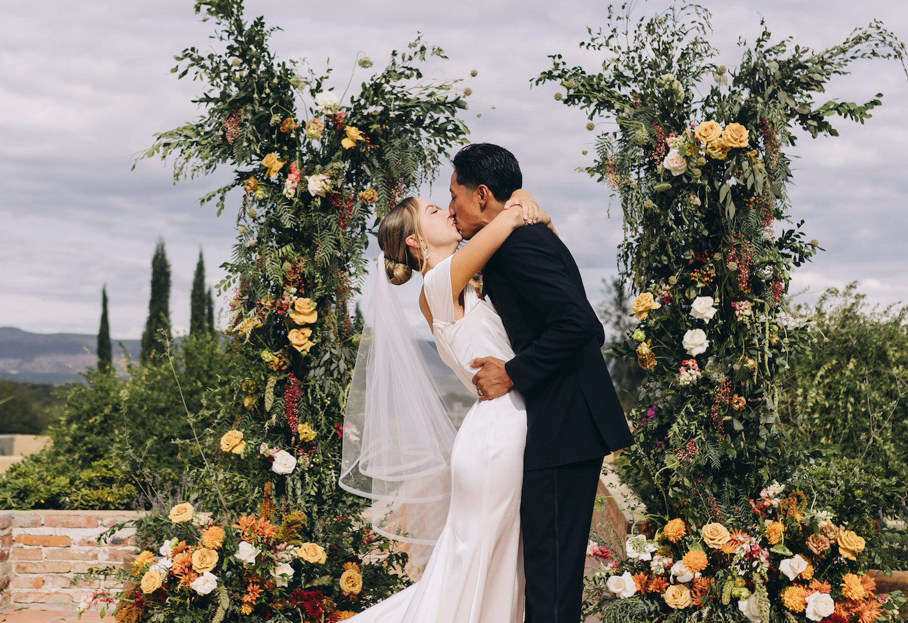 A bride and groom share a kiss at their wedding ceremony, standing between two floral arches, with greenery and mountains in the background.