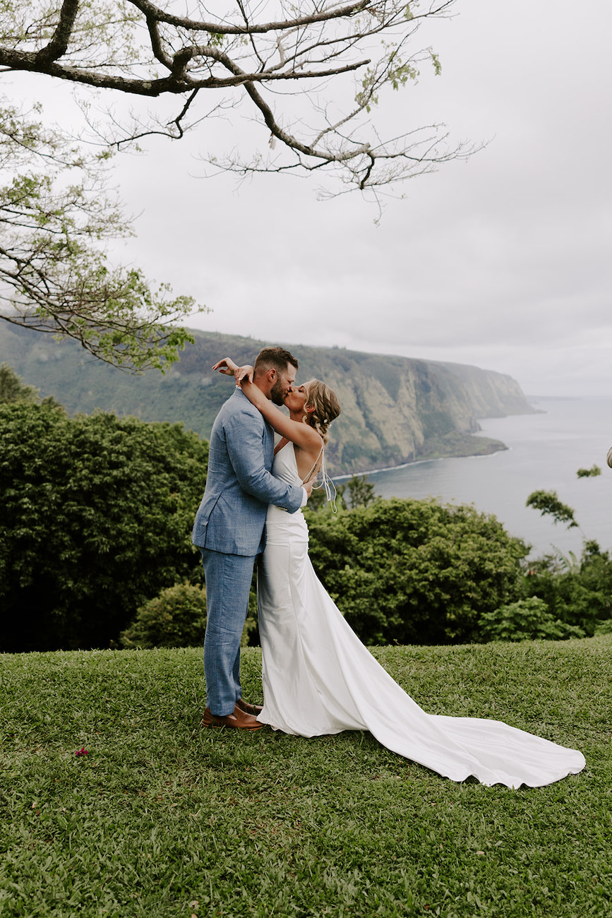 A couple in wedding attire embraces on a grassy area with a scenic backdrop of mountains and ocean. The groom wears a light blue suit, and the bride wears a white dress with a long train.