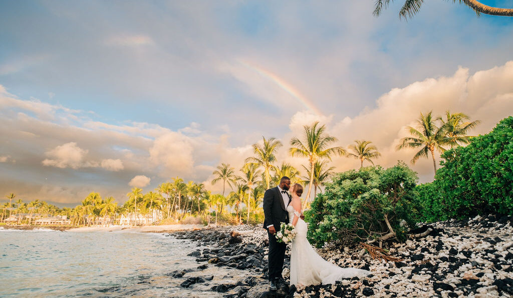 A bride and groom stand on a rocky beach with palm trees and a rainbow in the background. The bride holds a bouquet, and they are dressed in formal wedding attire.