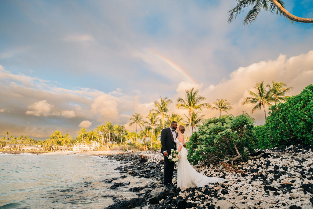 A bride and groom stand on a rocky beach with palm trees and a rainbow in the background. The bride holds a bouquet, and they are dressed in formal wedding attire.