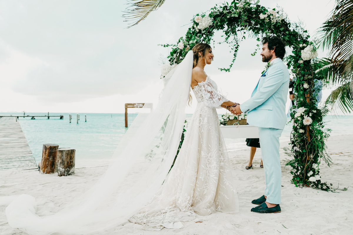 A couple stands holding hands under a floral arch at a beach wedding ceremony. The bride wears a white gown with a long train, and the groom wears a light blue suit.