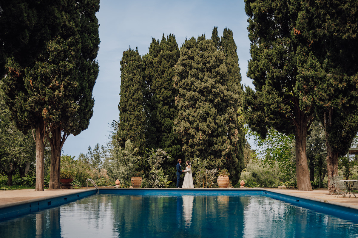 A couple stands near the end of a long, rectangular pool surrounded by tall, lush trees on a clear day.