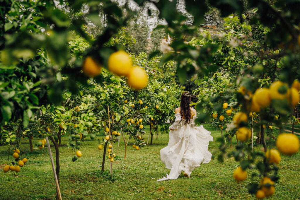 A person in a flowing white dress moves through a lemon orchard, surrounded by green foliage and yellow lemons.