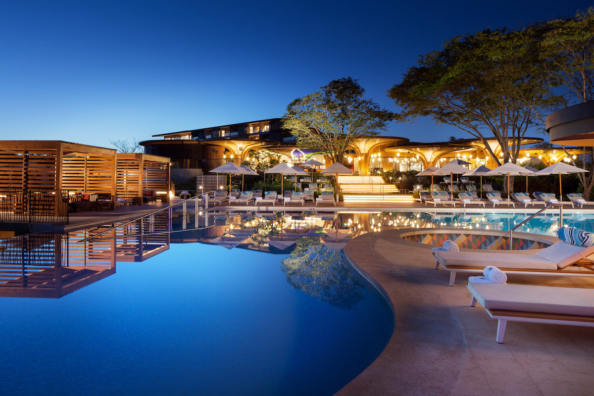 Luxurious outdoor pool area at a resort during evening, with lounge chairs, cabanas, umbrellas, and trees, all illuminated by warm lights. The sky is clear, transitioning from sunset to night.