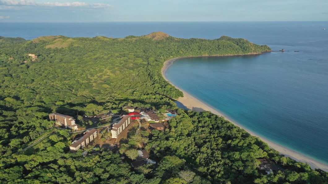 Aerial view of a lush, green forest surrounding a resort near a secluded beach with turquoise waters and a gently curving coastline under a clear sky.