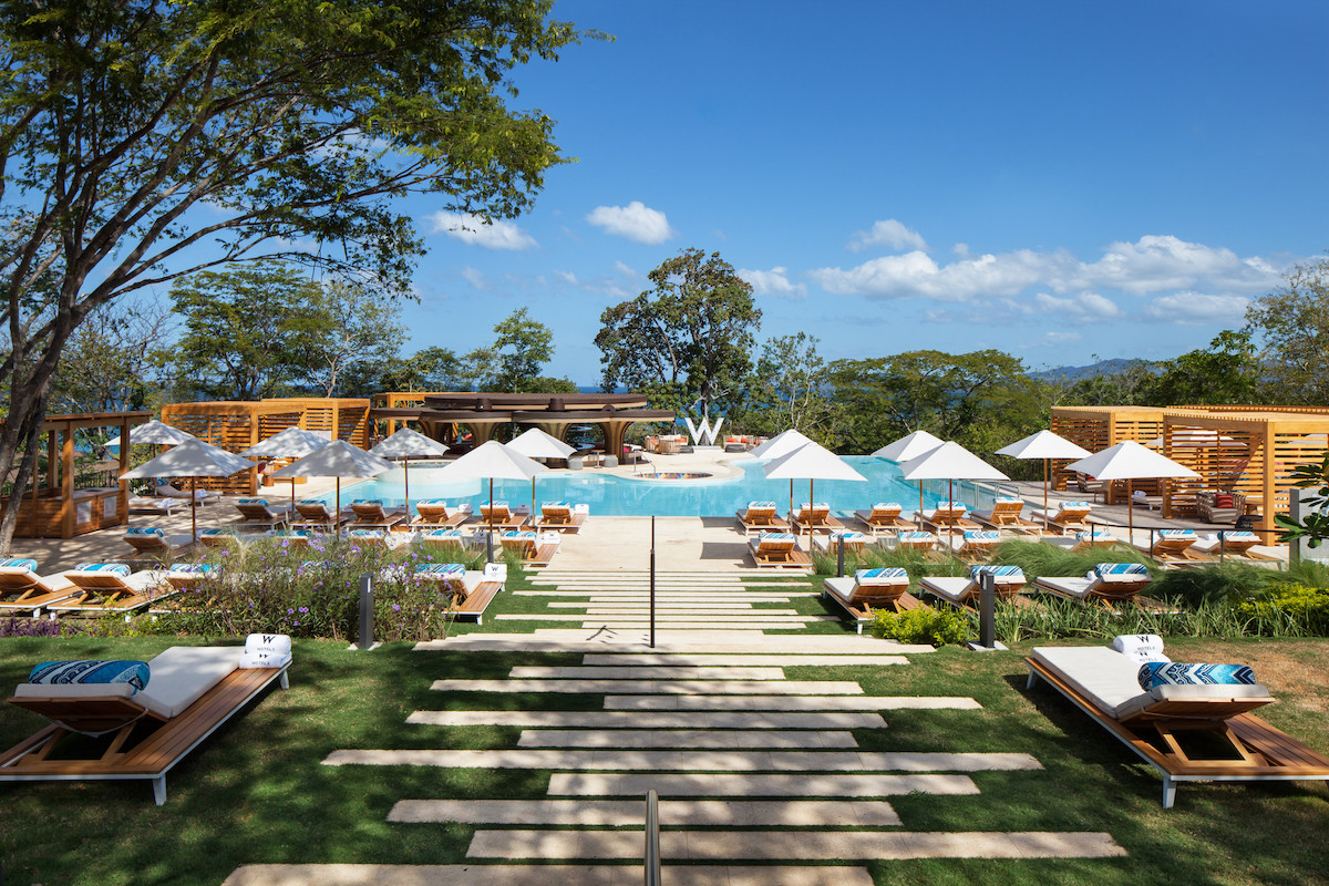 A resort pool area with lounge chairs, umbrellas, and cabanas set amidst greenery and trees, under a clear blue sky.