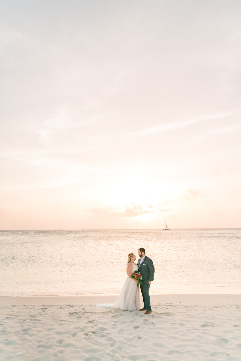 A couple in wedding attire stand on a beach at sunset, with a sailboat in the distance on the horizon and a calm sea.