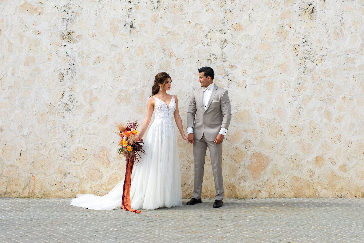 A bride and groom stand holding hands, facing slightly towards each other, against a textured stone wall. The bride holds a colorful bouquet with long ribbons.