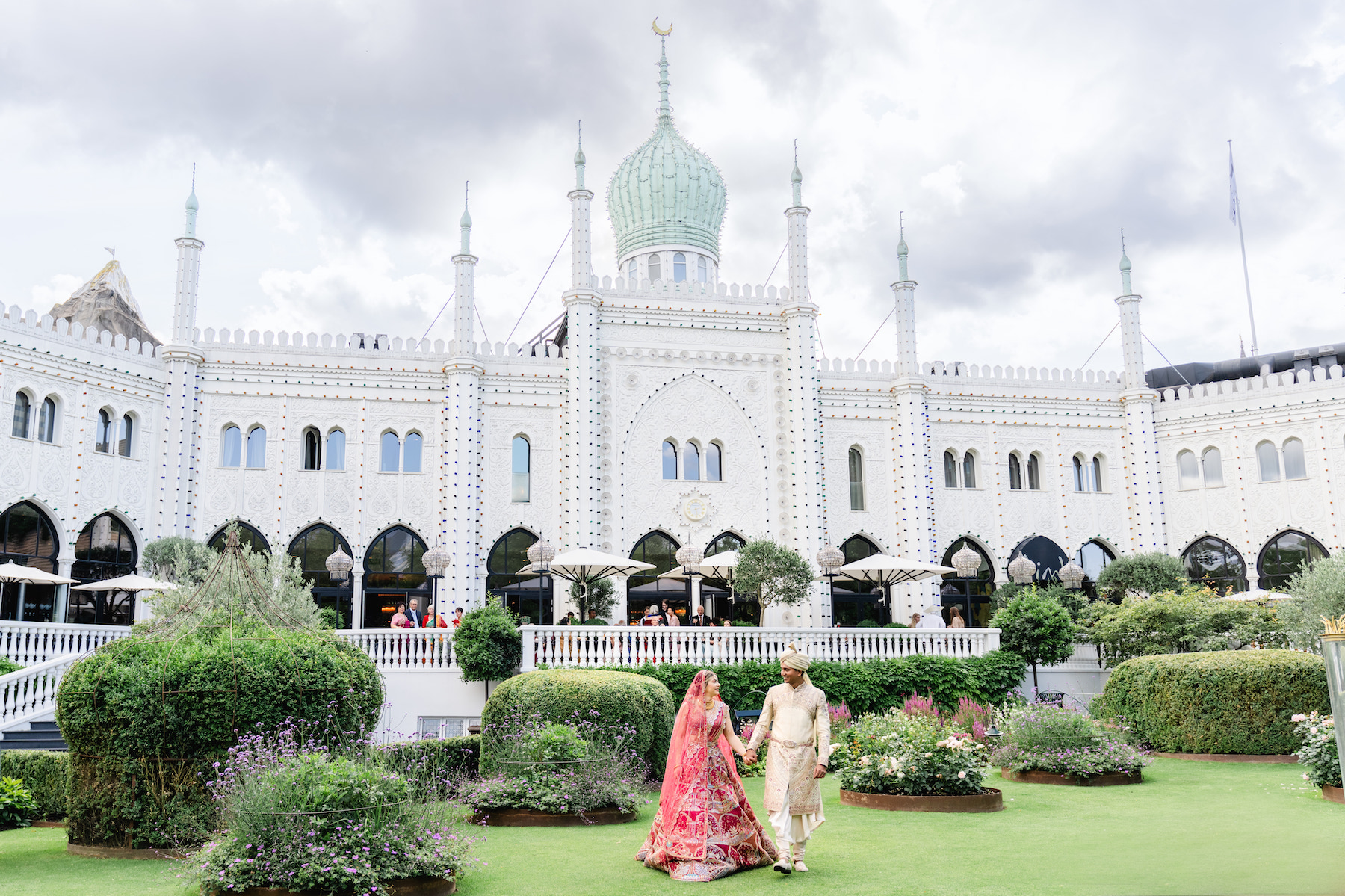 A couple in traditional attire walk hand in hand across a manicured garden in front of a grand, ornate building with a large central dome and arches.