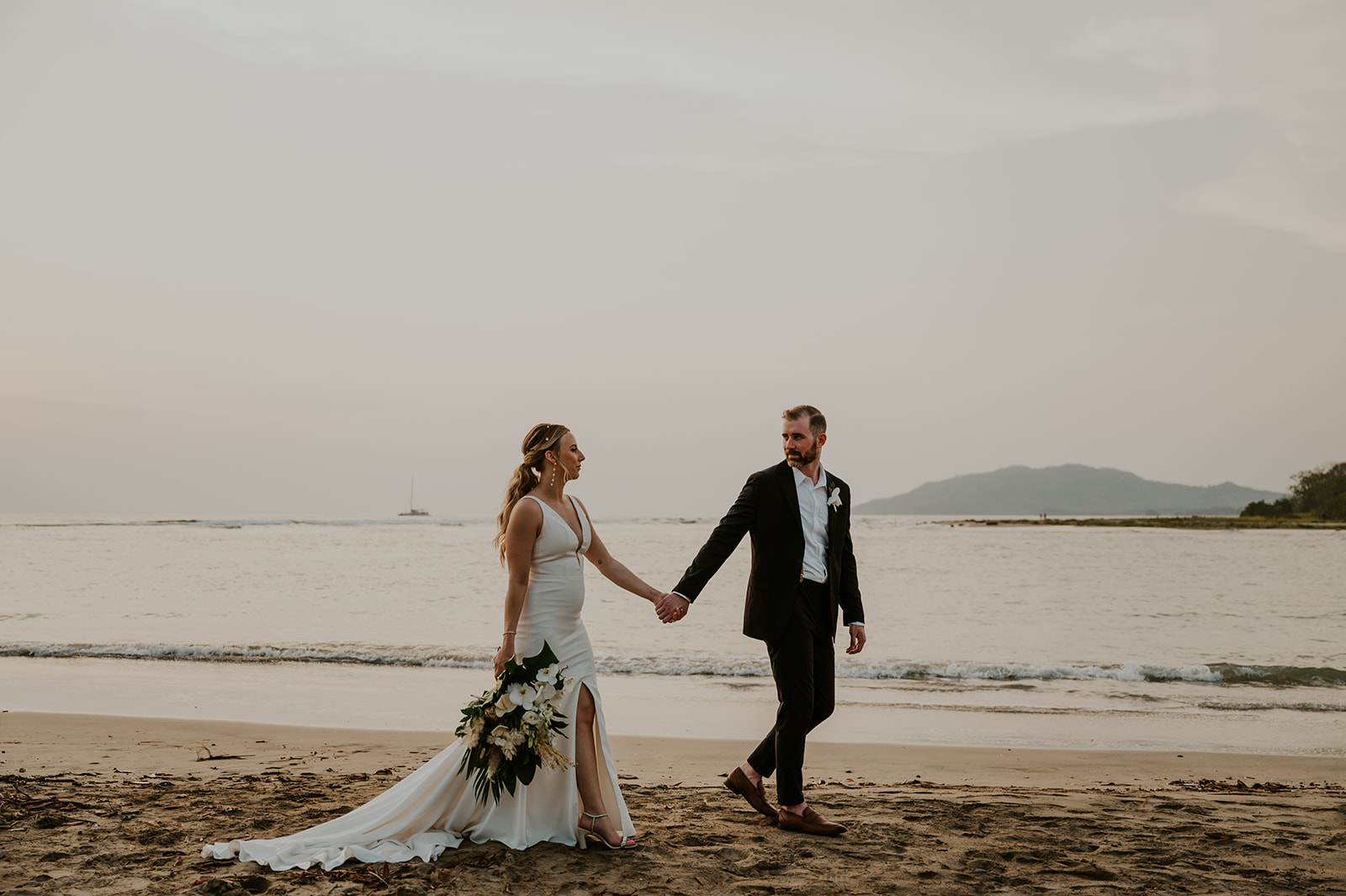 A couple in formal attire walks hand in hand on a sandy beach with the sea and distant mountains in the background. The bride holds a bouquet in her left hand.