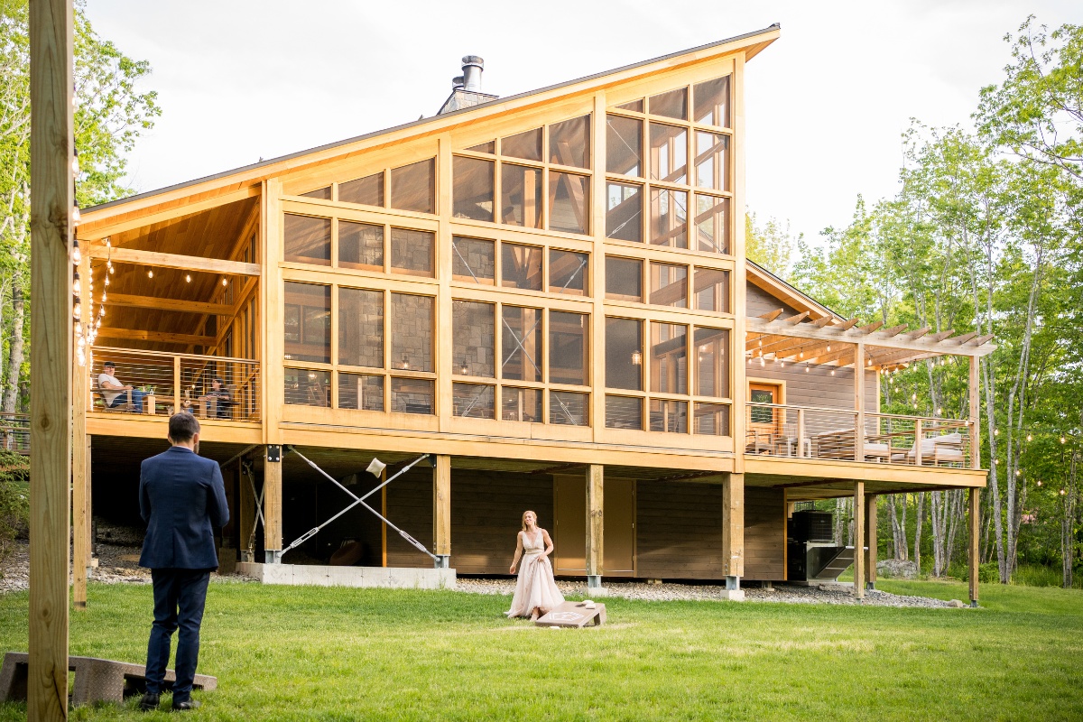 A person in a wedding dress walks towards another person in a suit standing on a grassy area in front of a large modern wooden house with extensive glass windows.