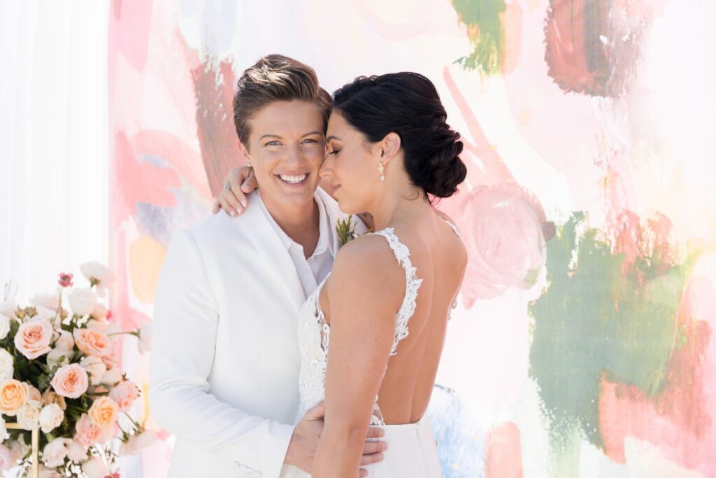 Two people in white wedding attire pose closely in front of a colorful backdrop, one smiling and the other resting their head on their shoulder. Floral arrangements are visible on the left side.