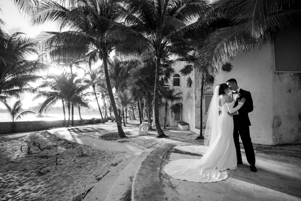 A bride and groom embrace under palm trees beside a sandy beach near a building, with sunlight filtering through the trees in this black and white photo.