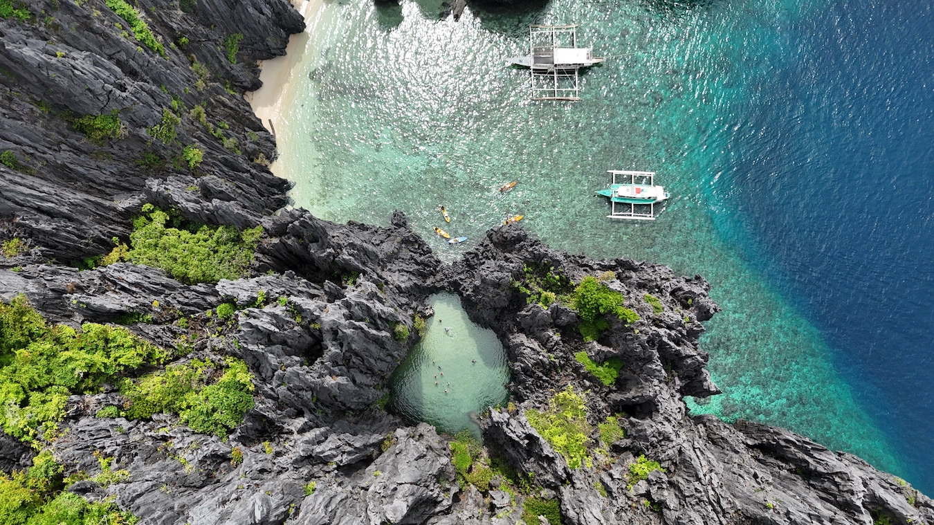 Aerial view of a rocky coastline with clear turquoise water, two white boats, and several kayaks near the shore and lagoon.