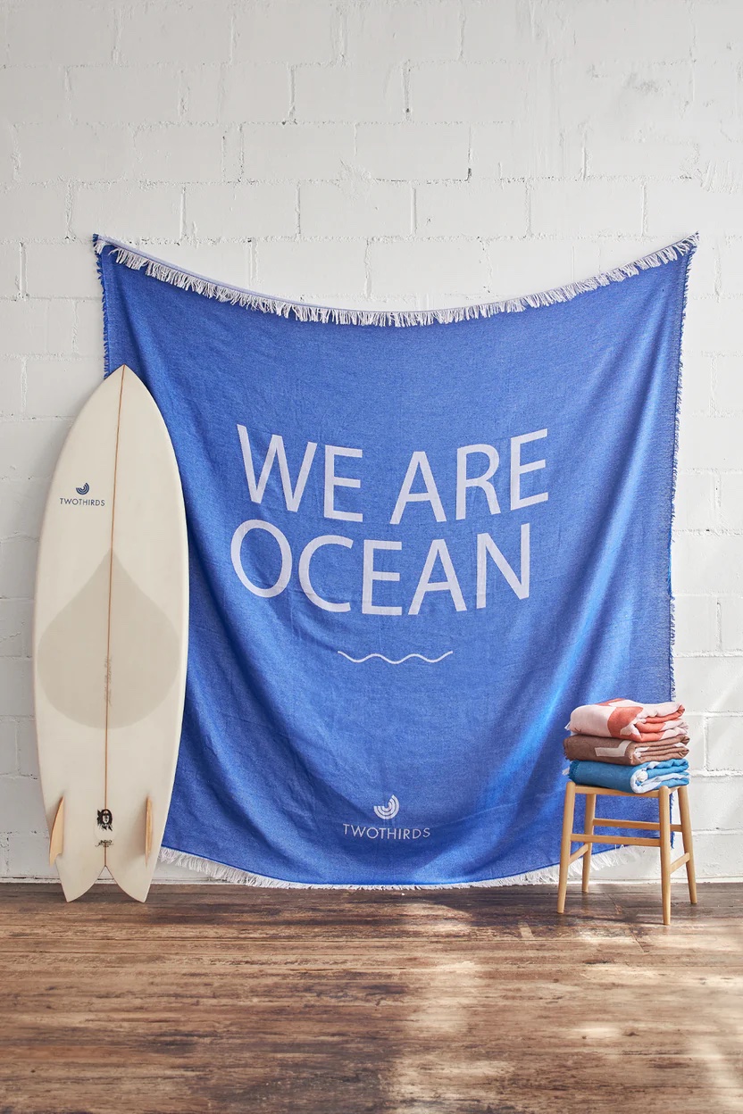 A blue tapestry with "WE ARE OCEAN" text hangs on a white brick wall. A surfboard leans against the wall, and a small stool with folded towels sits beside it.