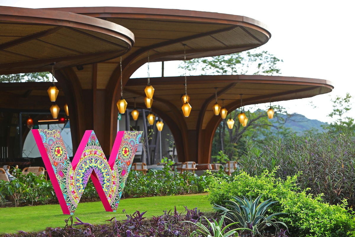 Outdoor seating area of a modern, circular-roofed structure with hanging lights, adjacent to greenery and a large decorative letter "W."